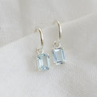 A pair of silver huggie style hoop earrings with rectangular blue topaz gemstone drops.