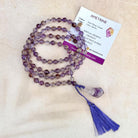 Purple amethyst gemstone beaded mala prayer necklace with a blue cord tassel, displayed next to an 'AMETRINE' product card on a light beige textured background.