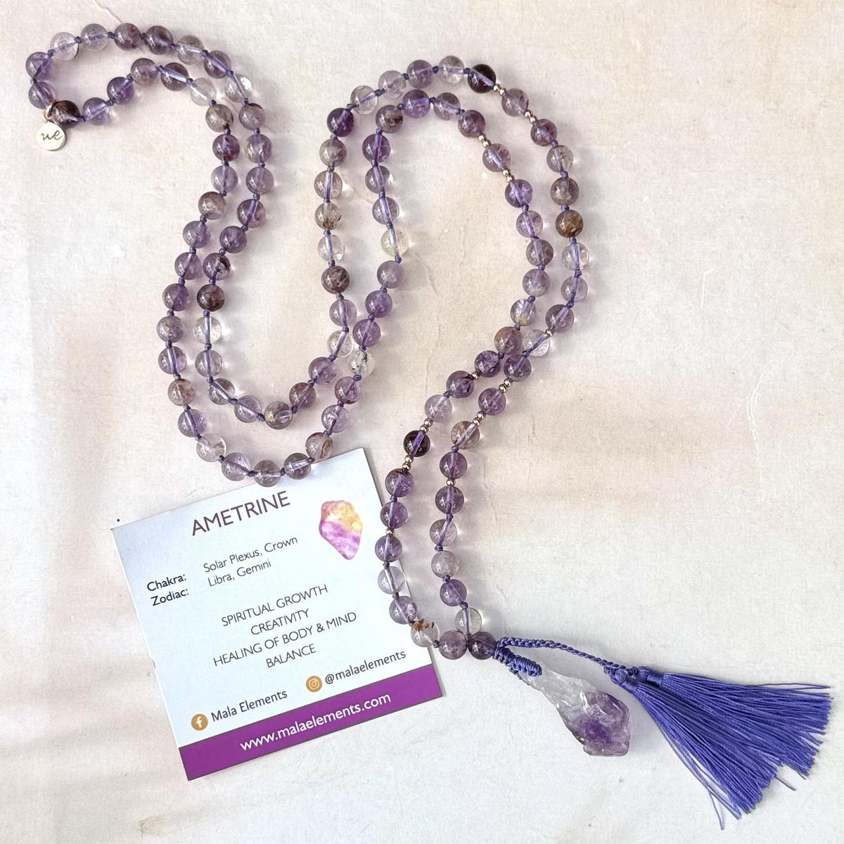 A purple ametrine bead mala necklace with a tassel and a white product tag, displayed on a light beige textured background.