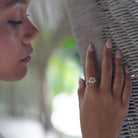 A close-up lifestyle shot of a woman's hand resting against a woven wicker wall, wearing a silver ring with a sparkling round solitaire gemstone.